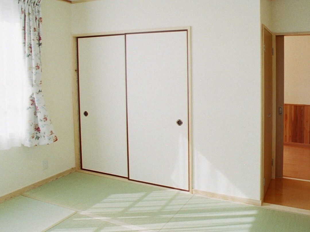 押し入れが付いた畳敷きの部屋にはレースカーテンとカーテンが付いたおおきな窓がある。家具は置かれていない。
