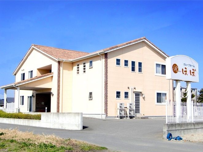 明るいベージュ系の2階建ての建物には三角屋根が付き、玄関には屋根がある。敷地内には駐車スペースもある。