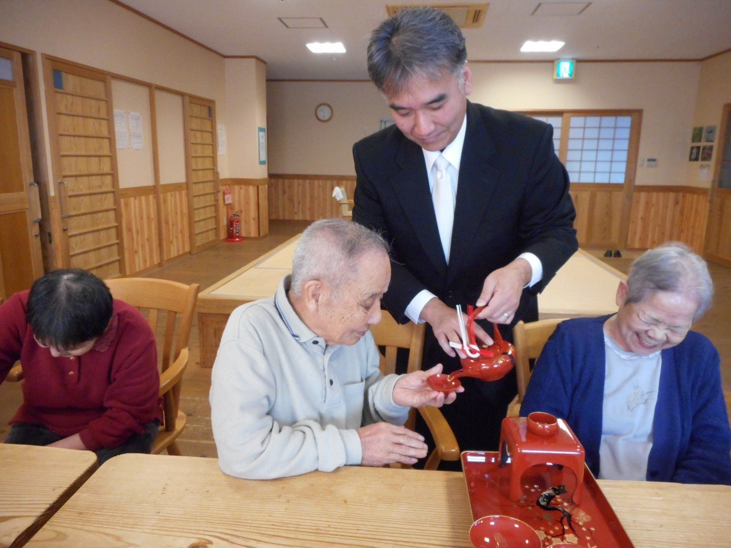 白いネクタイに黒スーツを身につけた男性スタッフがテーブルについている入居者にお屠蘇をふるまっている様子。高齢男性が杯を持って入れてもらっている。
