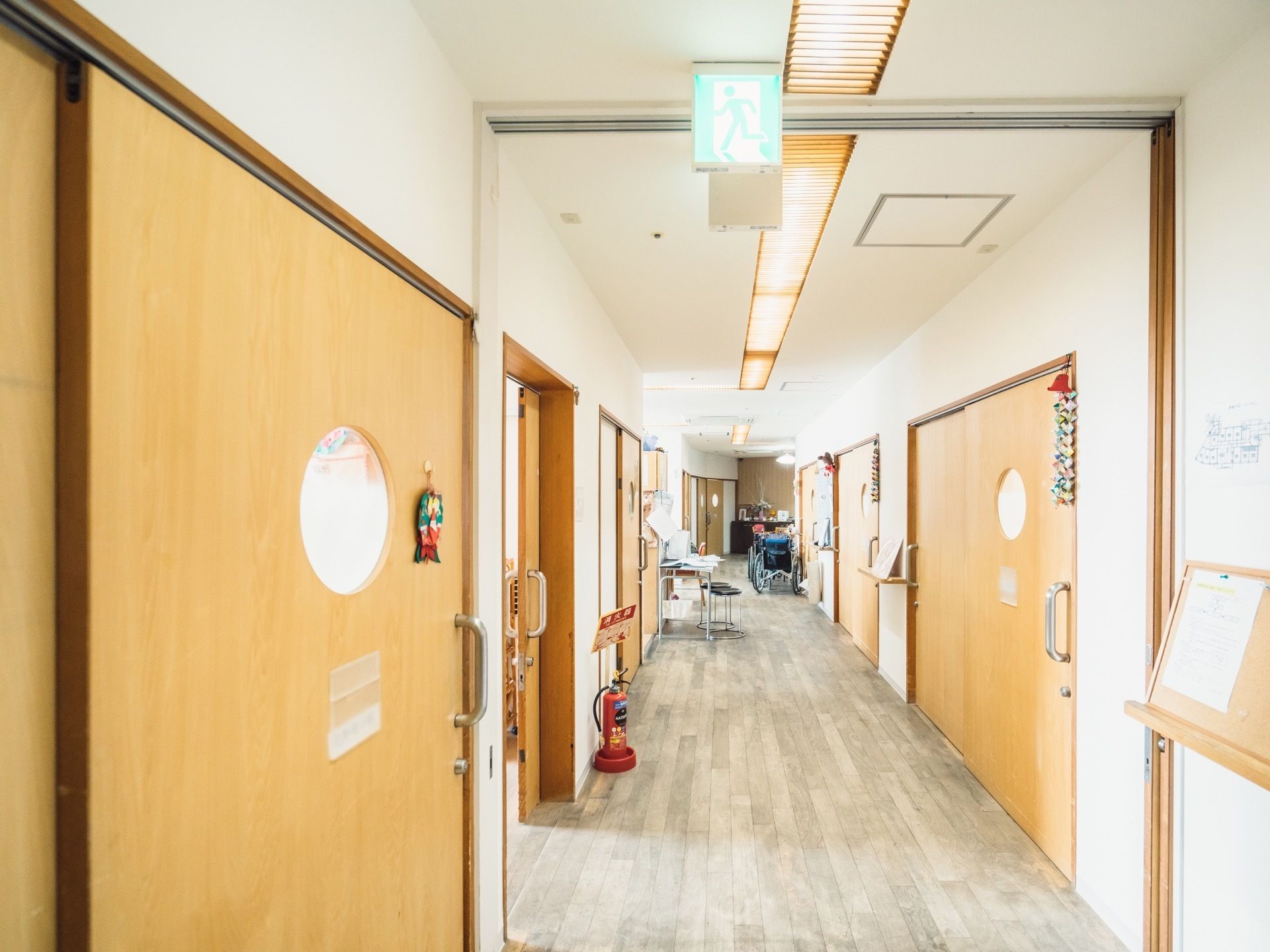 広々とした真っ直ぐな廊下である。左右に居室の扉が並んでおり、壁際に消火器が設置されている。奥に車椅子が置かれている。