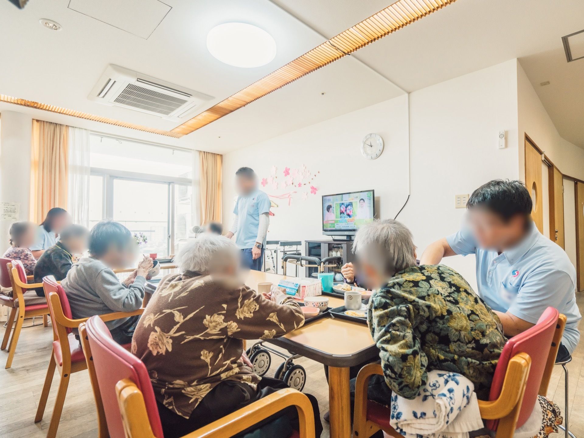 明るく開放的な食堂で、入居者様が揃ってお食事を召し上がっている。テレビが壁際に設置されている。スタッフが見守っている。