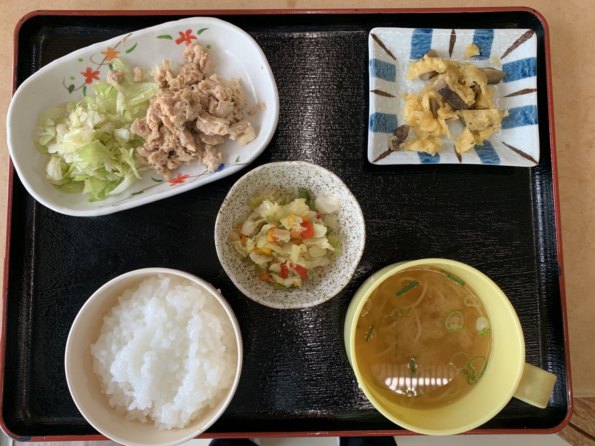 テーブルの上に黒いトレイが置かれ、ご飯茶碗やおかずのお皿が並べられている。木製の箸やスプーンが用意されている。