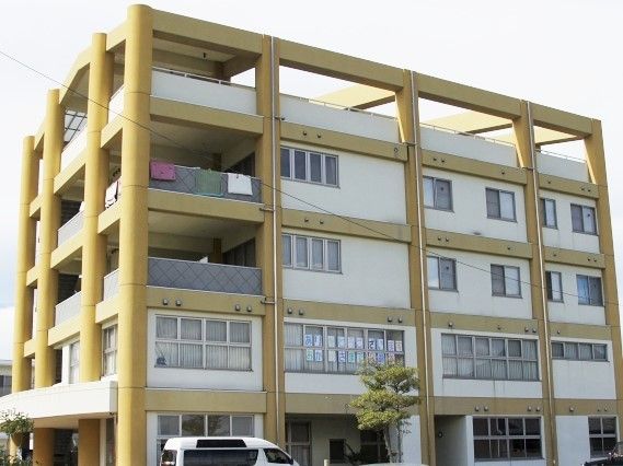 白とベージュの大きな建物。広いベランダも各フロアにつき、周辺には駐車場もある。