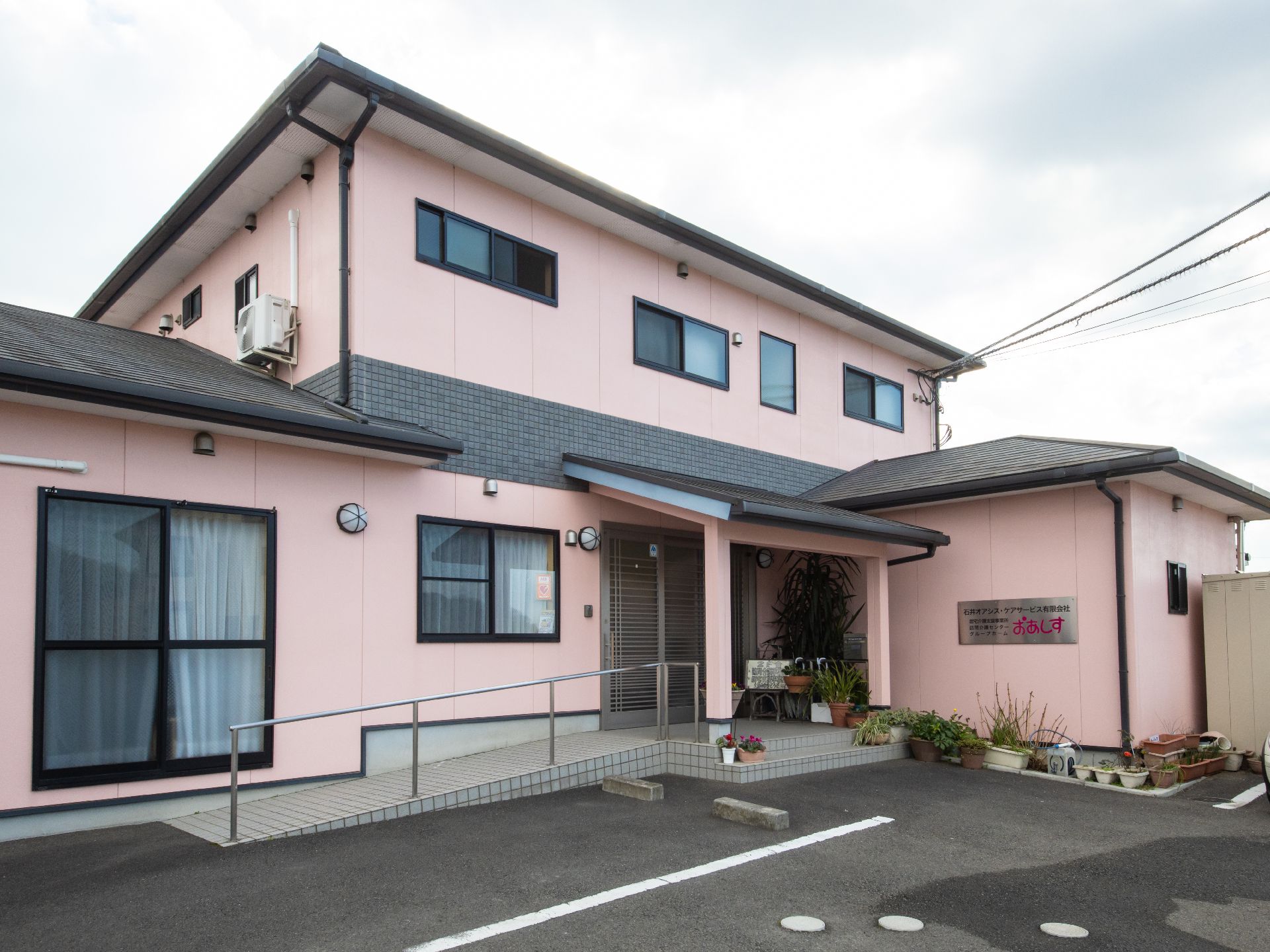 淡いピンク系の外壁が印象的な施設である。玄関横には、手すり付きのスロープがあり駐車場が完備されている。