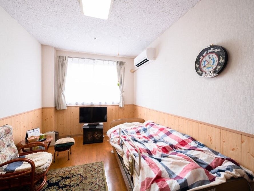 チェックの寝具付きのベッドやテレビ、椅子やテーブル、時計などがあるフローリングの部屋には大きな窓も付いている。
