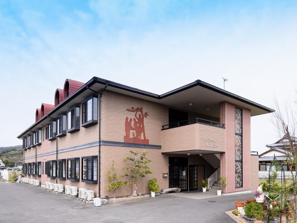 ブラウンの外壁の2階建ての建物。窓が多く付き、玄関部分が少し入り込んでいて建物の周辺には植物が植えられている。