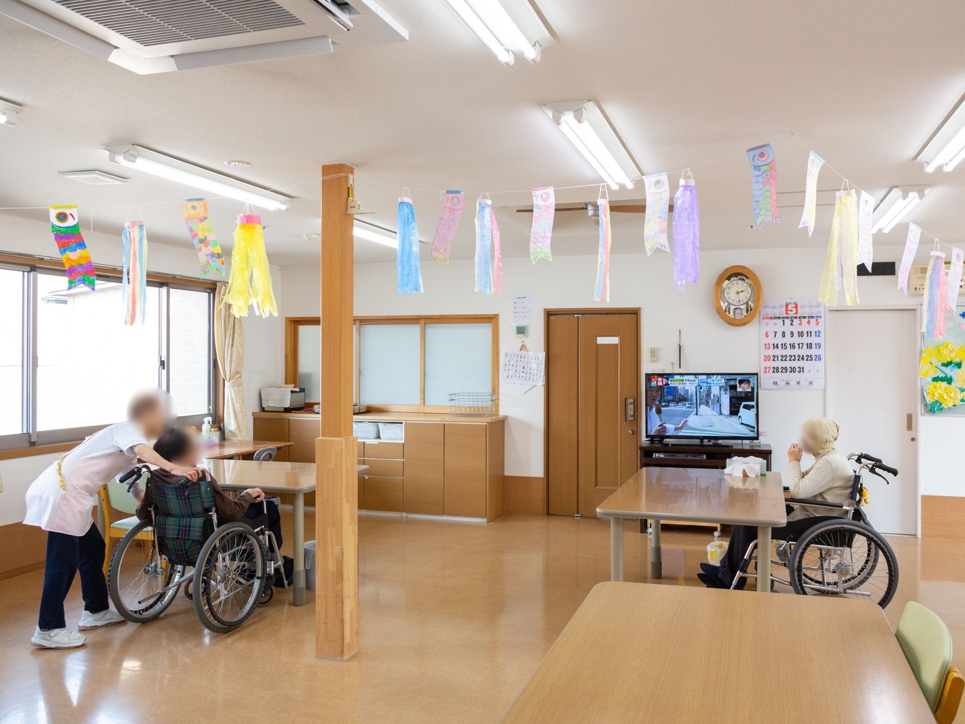 施設スタッフの介助を受けながら、入居者様がテレビを観賞されている。室内の天井にはロープがかかっており、小さな鯉のぼりがたくさん飾られている。