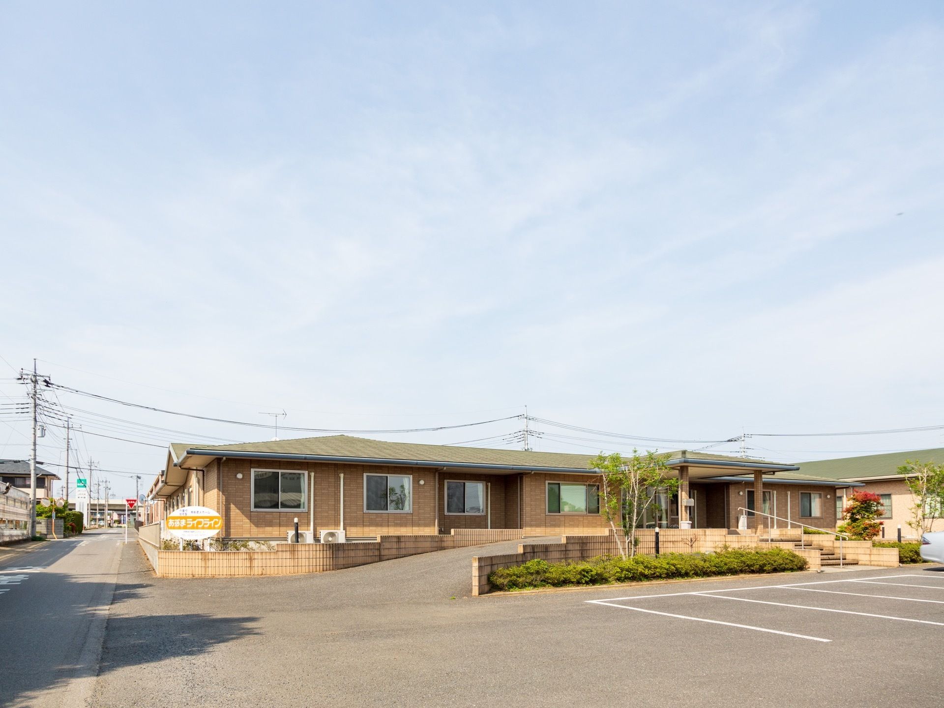 青空の下に平屋建ての施設が建っている。建物の屋根は緑色、外壁は茶色である。玄関まではスロープと階段が付いている。