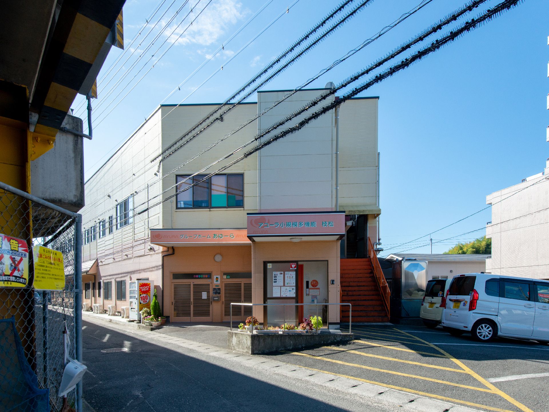 ブルーのエプロンを付けた若い女性スタッフが付き添い、屋外で笑顔で過ごしている車いすの高齢女性。