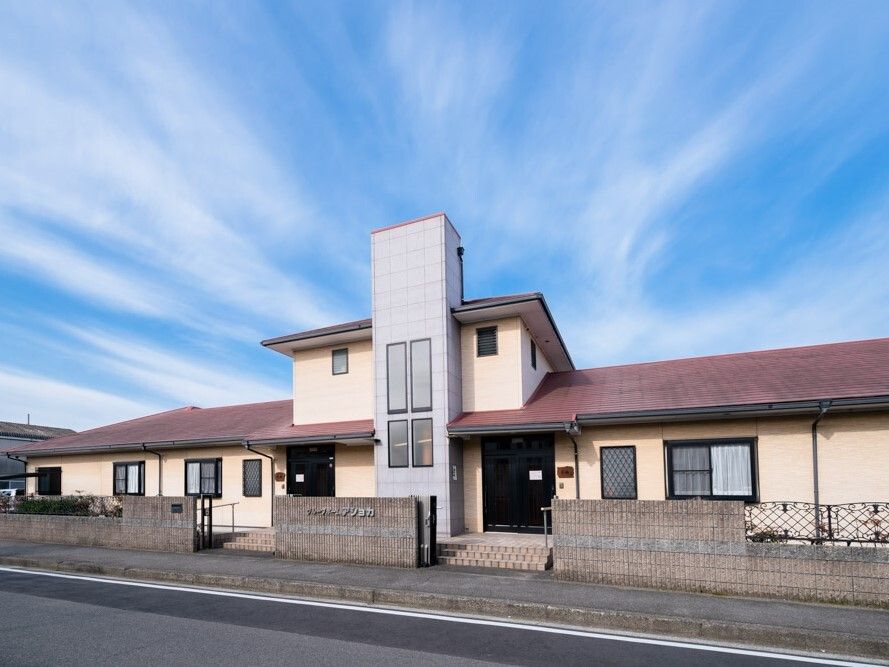 エンジ色の屋根が付いた2階建ての建物。敷地の周りにはブロック塀が建っていて青空が広がっている。