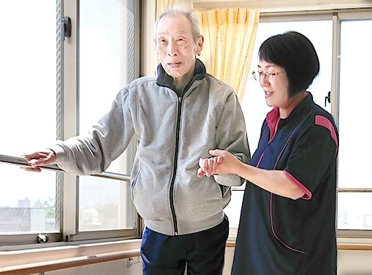 窓の手すりに手をかけ歩行訓練を行う背の高い男性入居のサポートに当たる女性スタッフ。