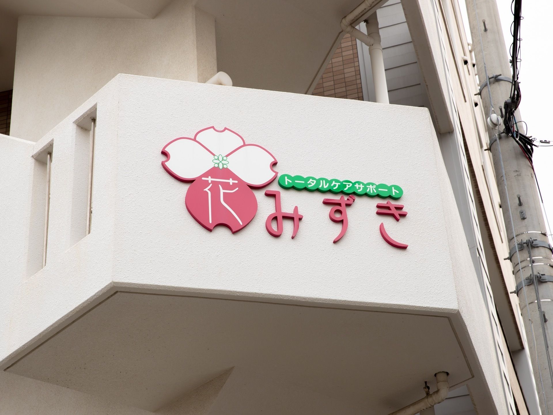 白を基調とした外壁に施設名が書かれている。花をモチーフとしたマークに、ピンク色で文字が書かれている。