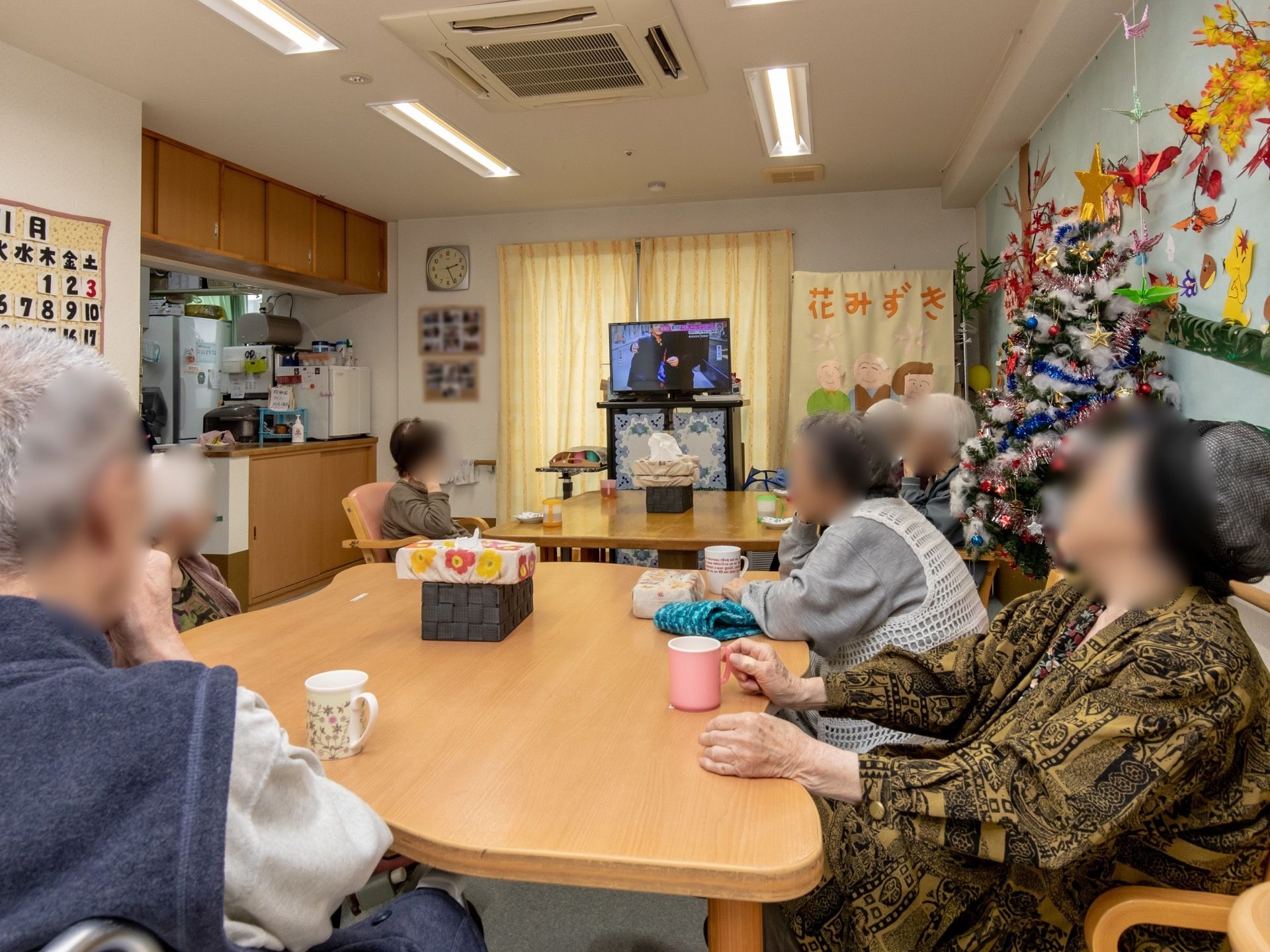 キッチンが併設されたリビングで、入居者様がテーブルを囲んで思い思いの時間を過ごしている。フロアにはツリーが飾られている。