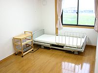 シンプルなフローリングの居室は窓が大きく、カーテンが設置され、ベッドなども置かれている。