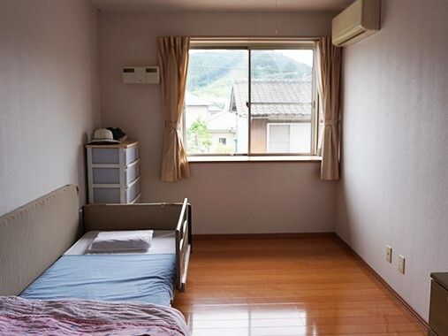 カーテンが付いた大きな窓が付いているフローリングの部屋には寝具付きのベッドが置かれ、エアコンもある。
