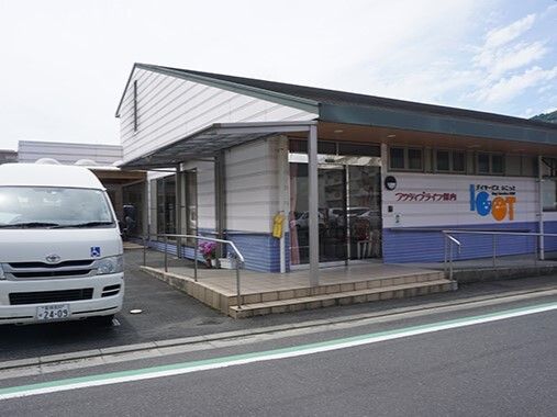 平屋建ての建物の外壁は白く、ホーム名が書かれている。敷地内には福祉車両も止まっている。