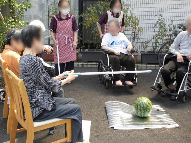 緑の多い屋外で外気浴を楽しんでいる車いすの高齢女性と付き添いの若い男性スタッフ。二人とも笑顔。