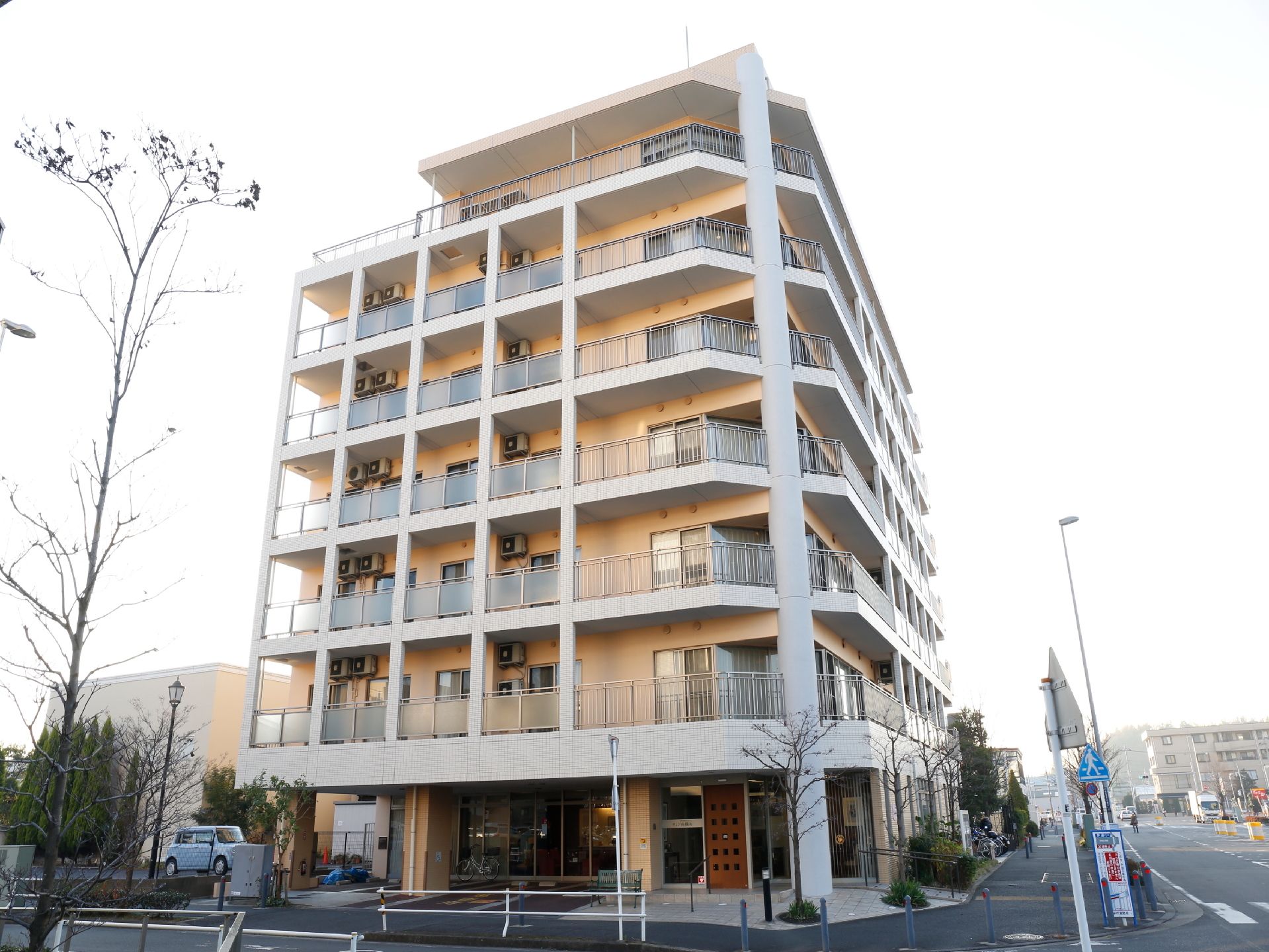 通りの角に立っているコンクリート造り7階建ての施設。建物は全体的にベージュ系の色で、入り口の茶色の扉が目立つ。