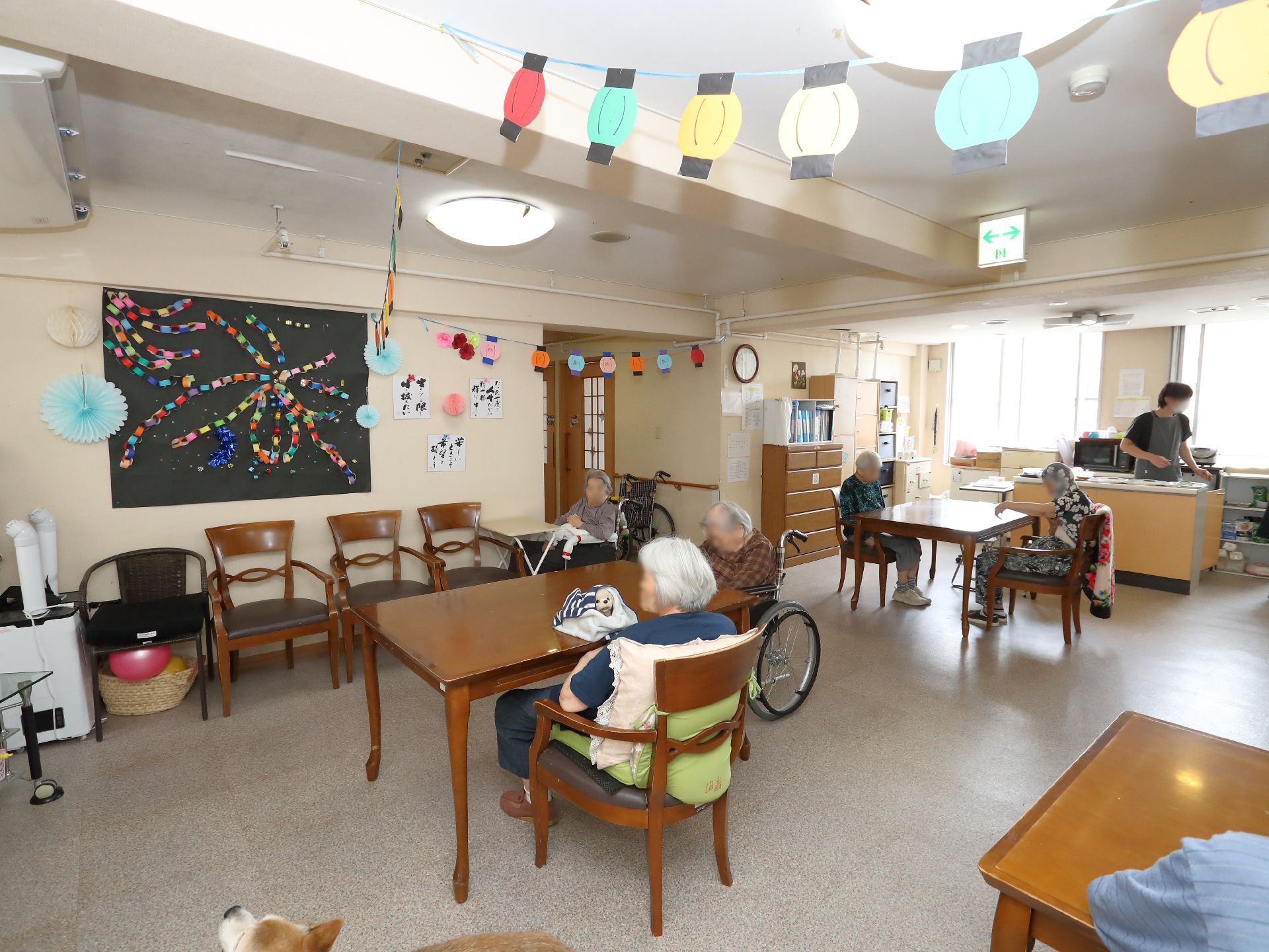 入居者様が周りに座る木製の机が置かれた白い壁の広間