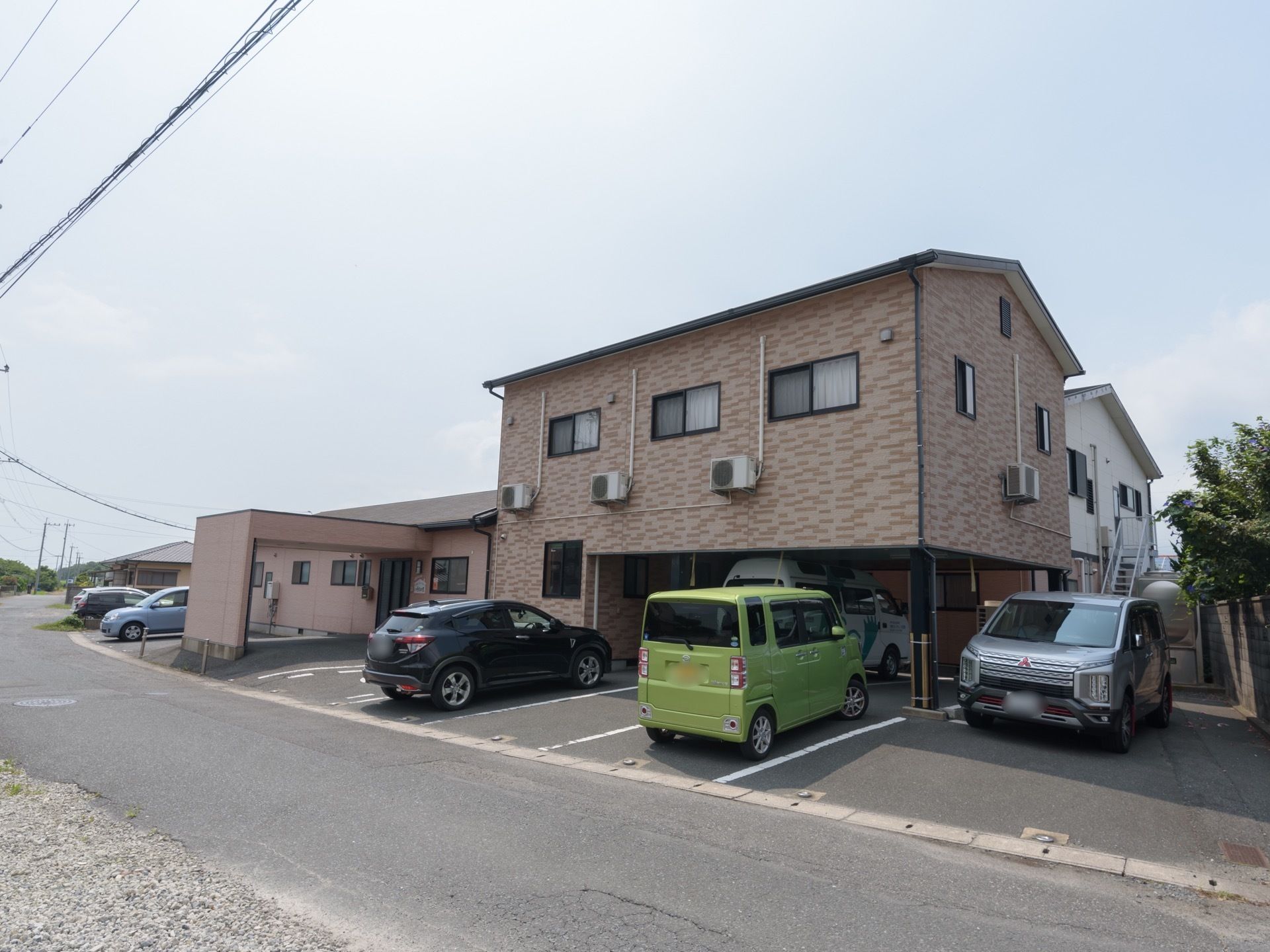 １階建てと２階建ての建物があり、その２つがつながって１つの建築物になっている。その建築物の周囲には駐車場があり、車が複数駐車されている。