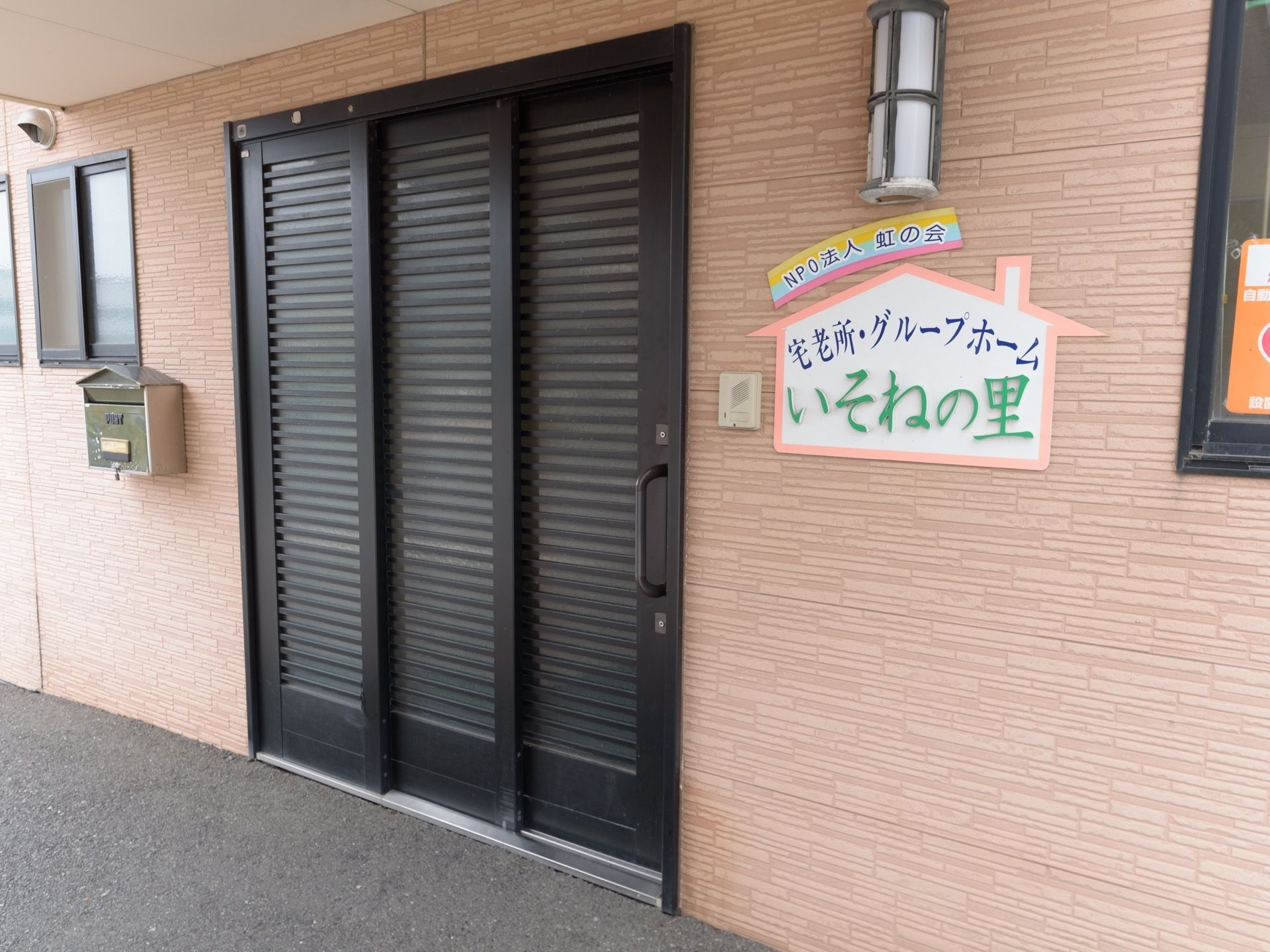 暖色系の外壁に黒色の引き戸の扉の玄関がある。玄関の横には、施設の名称が書かれたプレートが貼られている。