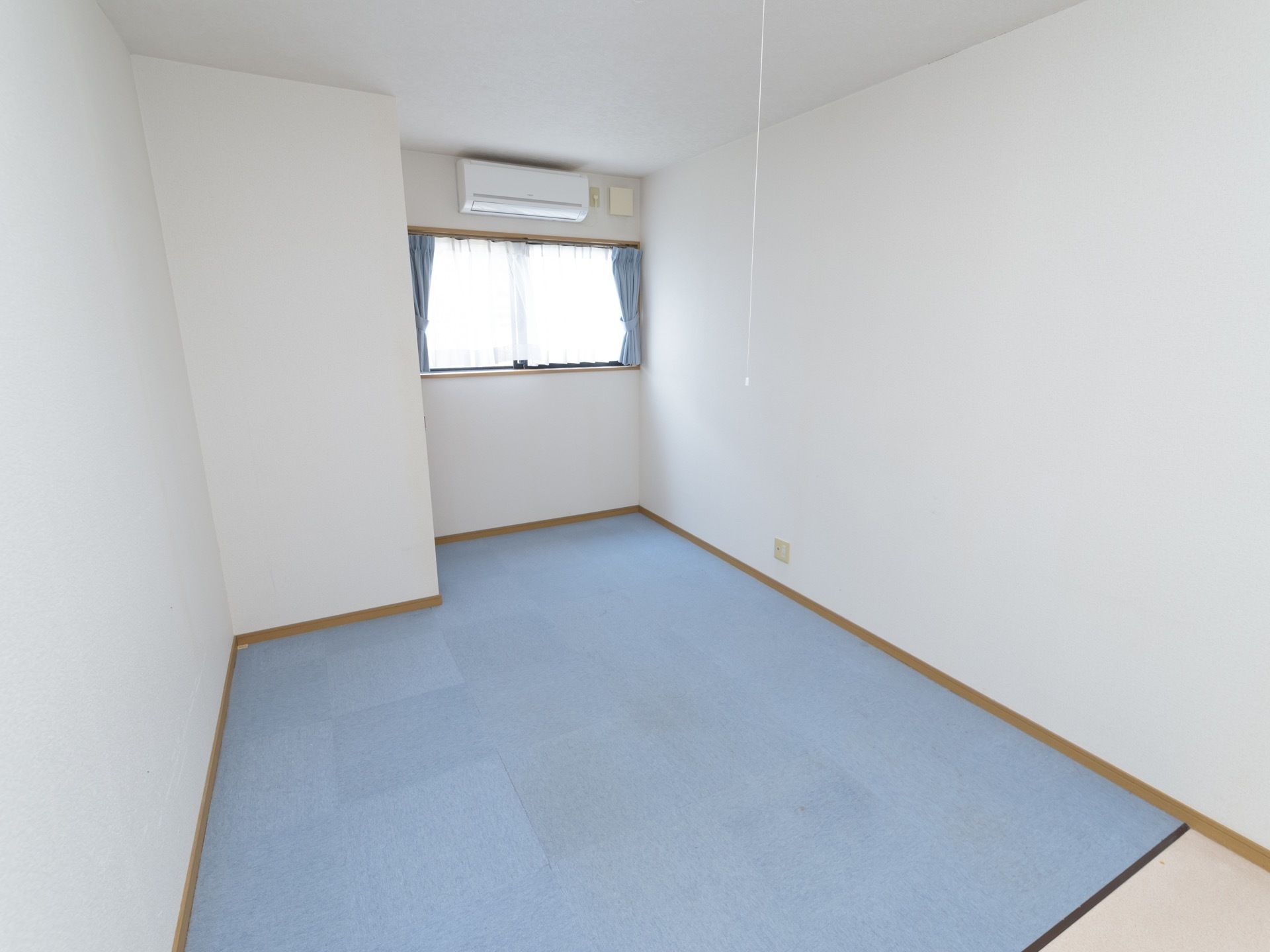 洋風の居室。床には青色の敷物が敷かれている。部屋の奥には窓があり、窓の上にはエアコンも設置されている。