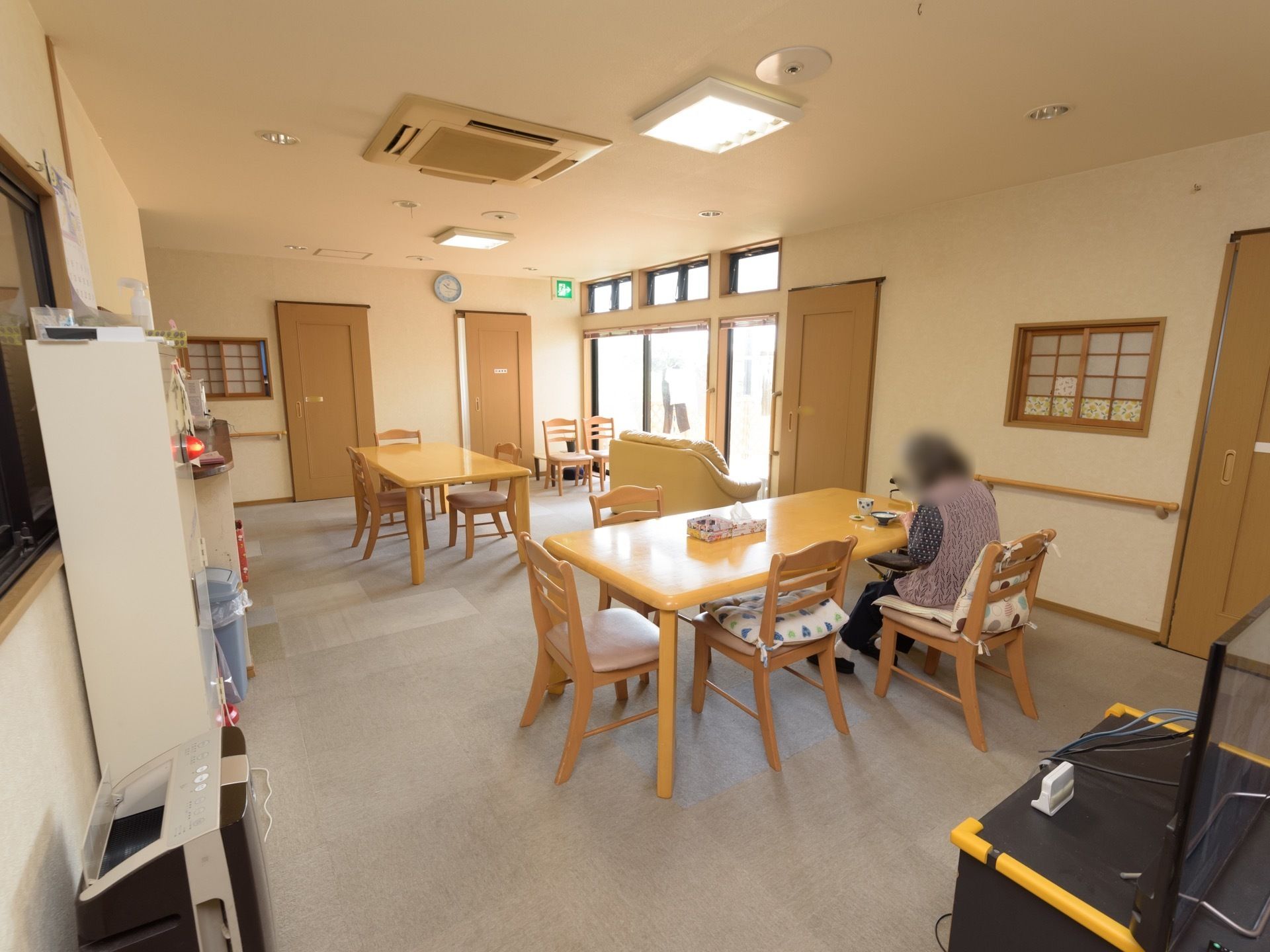 複数のテーブルと椅子のセットのほか、ソファなどが置かれた共有スペース。壁には手すりが備え付けられ、壁掛け時計なども設置されている。