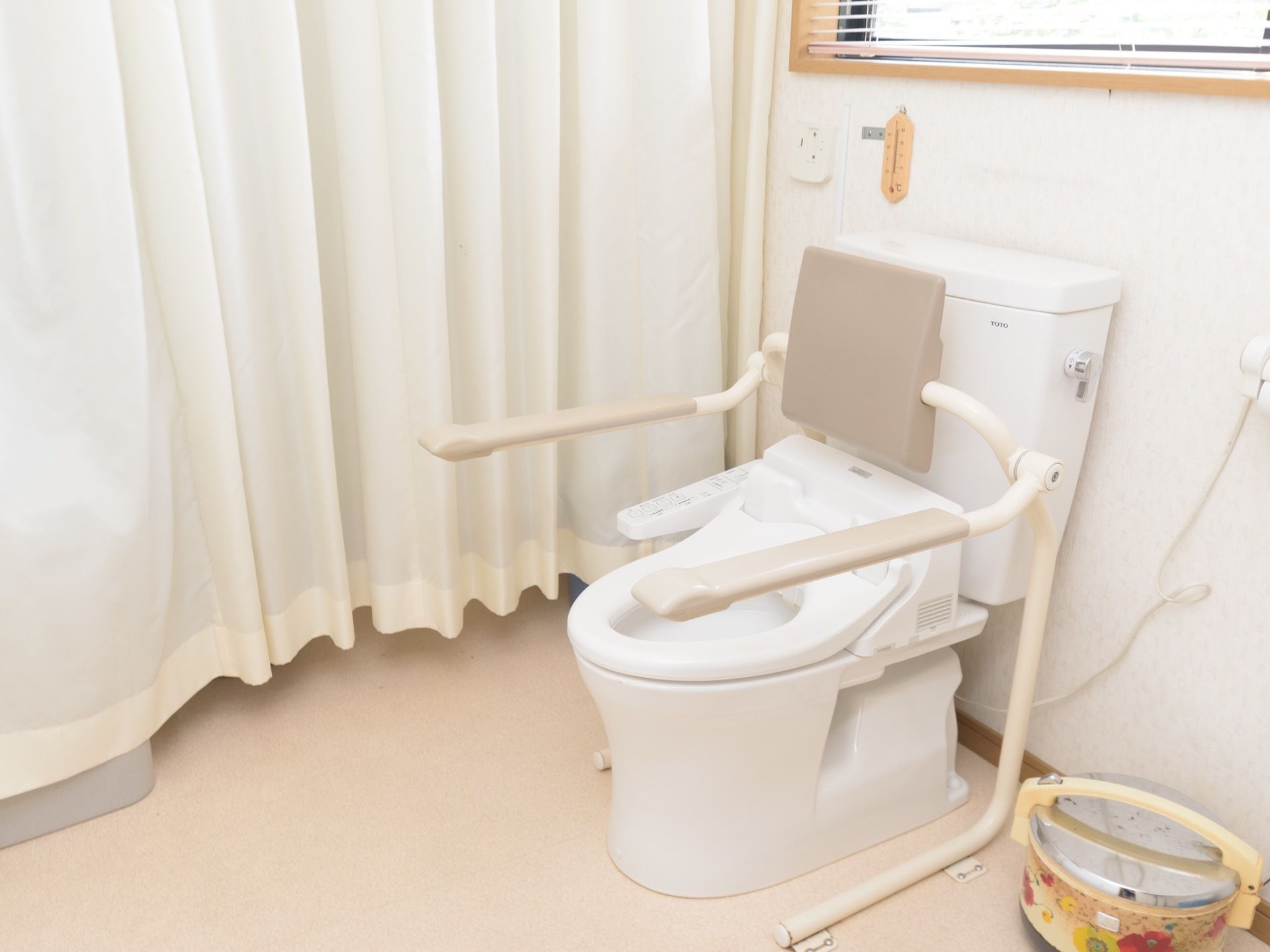 白いカーテンで仕切られたトイレの個室。ひじ掛けや温水洗浄便座などの設置された、洋式の便器が１つ備え付けられている。
