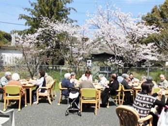 園庭にテーブルを並べ、桜を見ながら食事を楽しんでいる入居者とスタッフ。