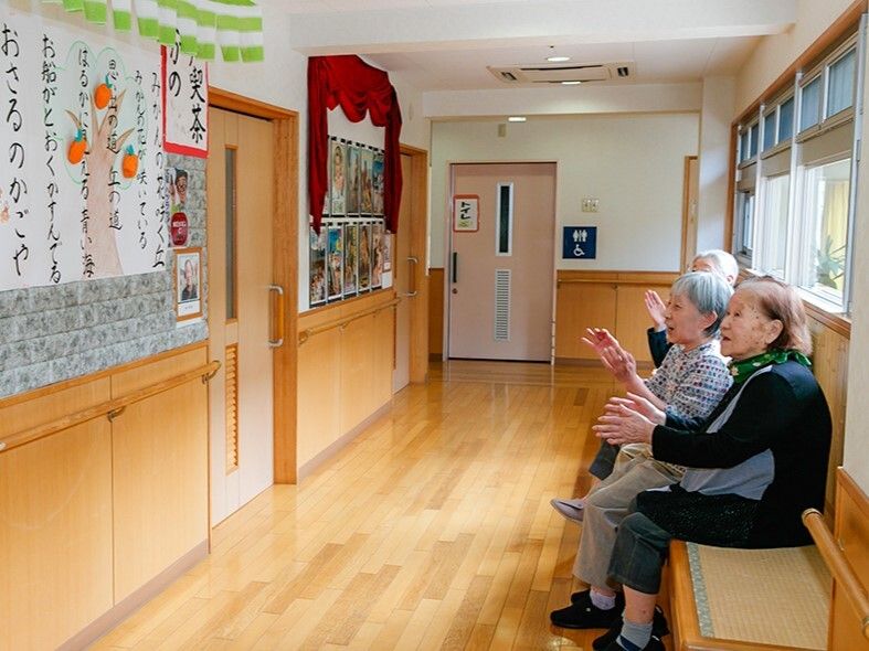 廊下のベンチに座って、壁に貼られた歌詞を見ながら、手拍子をして歌っている高齢女性たち。