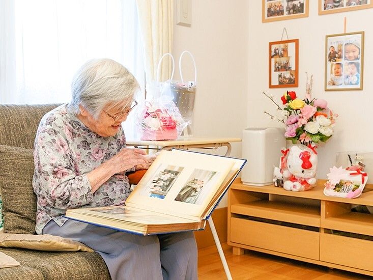 ソファに座って笑顔でアルバムをめくっている眼鏡をかけた高齢女性。周りには花やぬいぐるみがある。