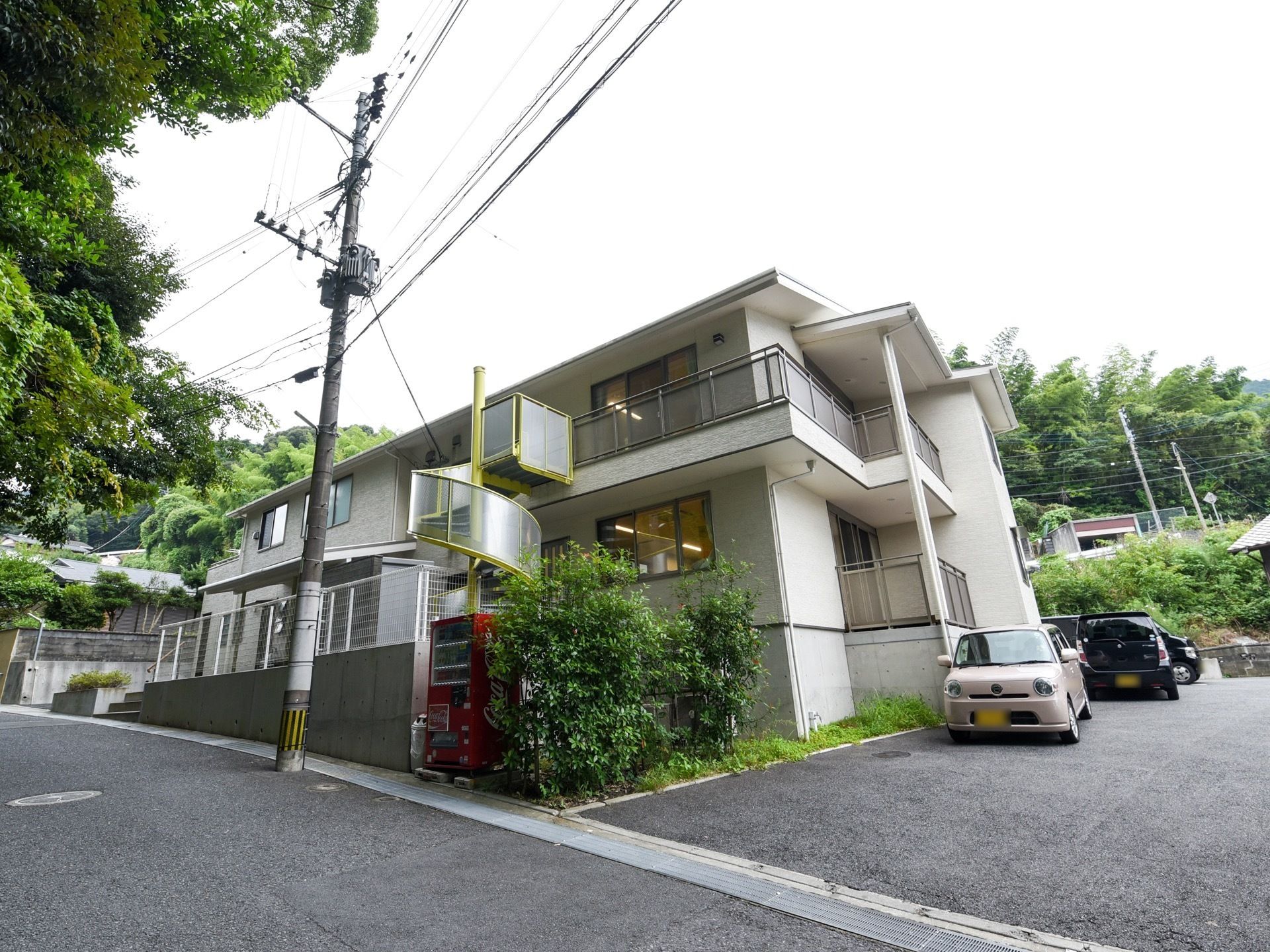 施設はらせん階段が印象的な２階建の白い建物である。建物横に駐車場が設けられている。植え込みの側に自販機が設置されている。