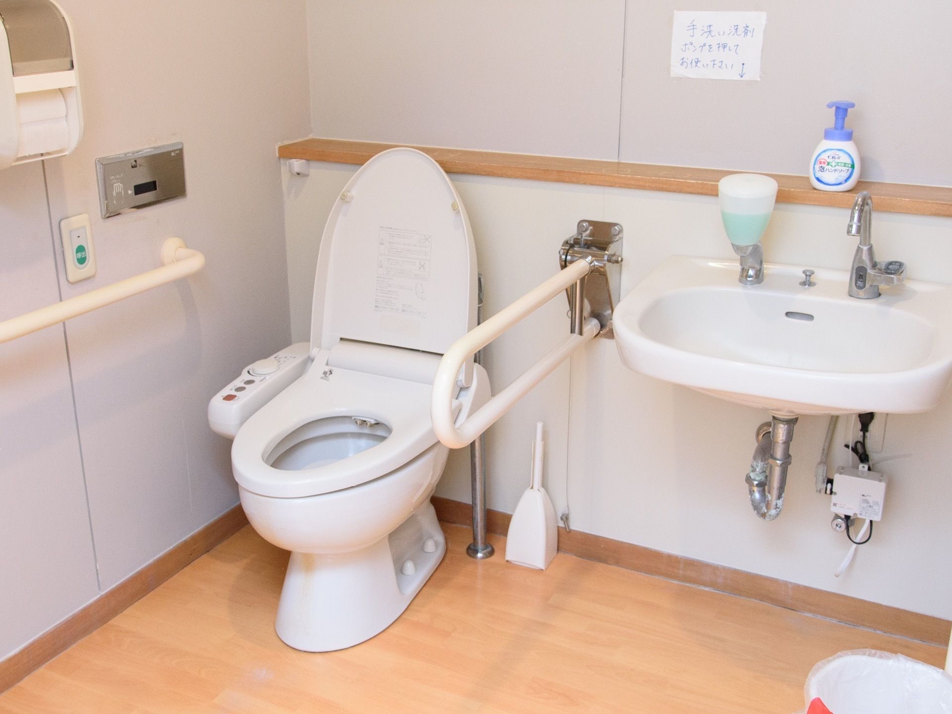 温水洗浄便座が採用された明るいトイレである。便器の両側に手すりがある。壁には呼び出しボタンがある。
