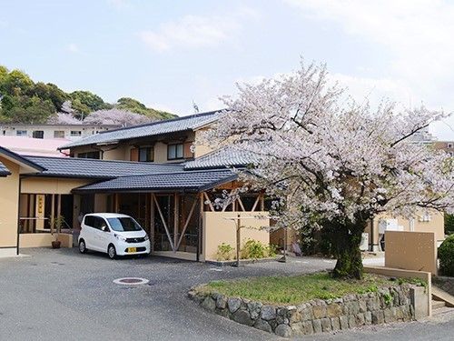 二階建ての一軒家風の建物が写っている写真。建物の前に駐車場と桜の木がある様子