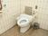 白いタイル張りの壁のあるトイレである。壁にはナースコールがあり、固定の手すりも見える。また、便器の両脇には可動式の手すりがある。