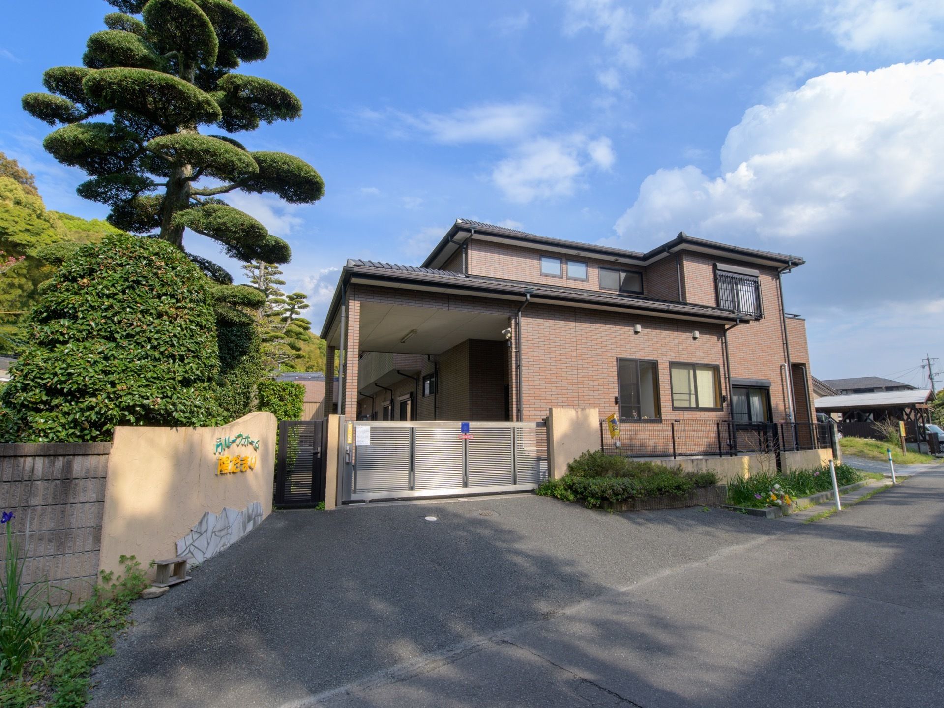 閑静な住宅街の中にあるグループホームで、周辺には高い建物がない。玄関には門が設置されており、塀に施設名が書かれている。