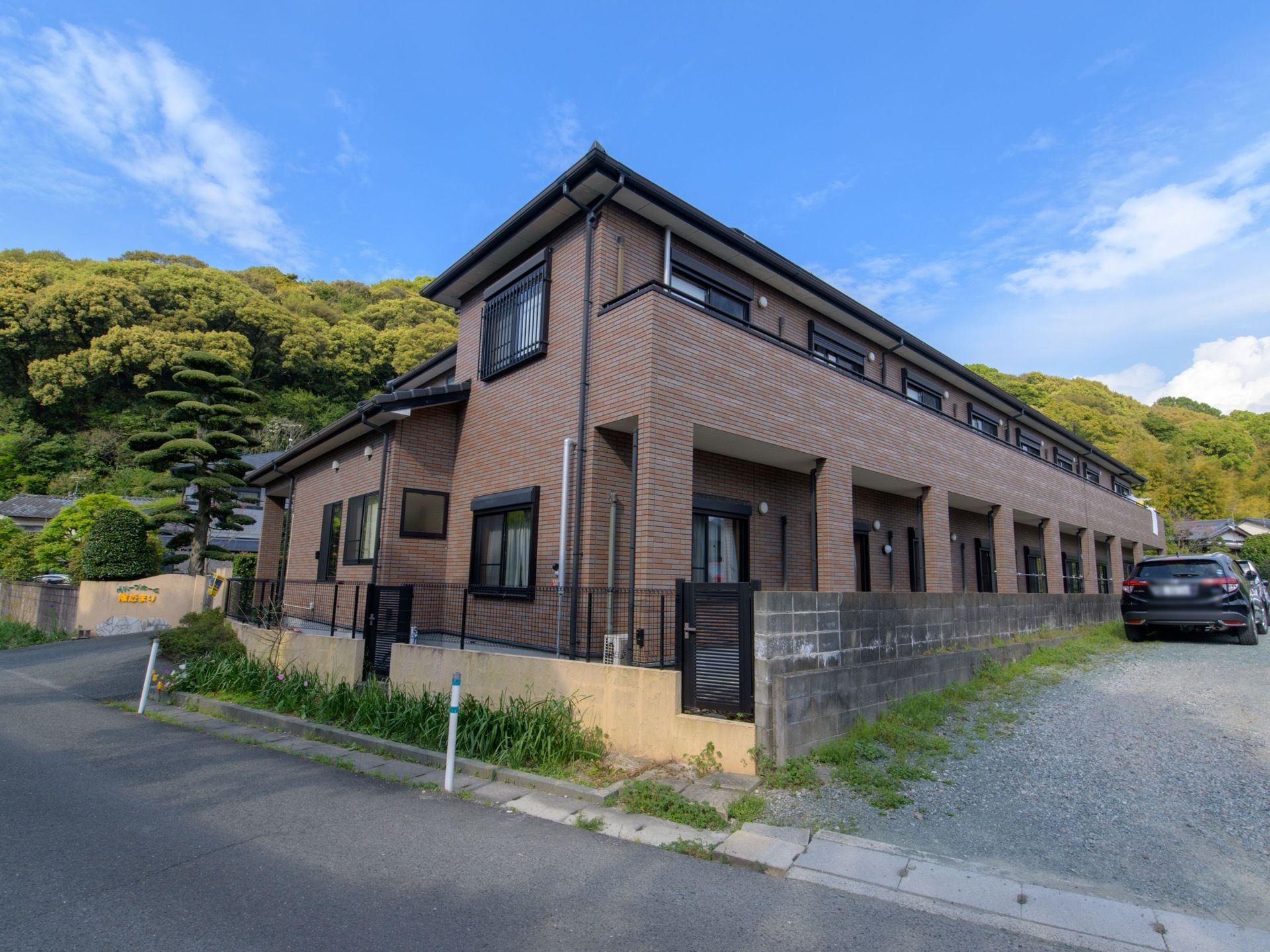 ブラウンを基調とした２階建ての建物で、横に駐車スペースが設けられている。周辺は緑豊かな山に囲まれている。