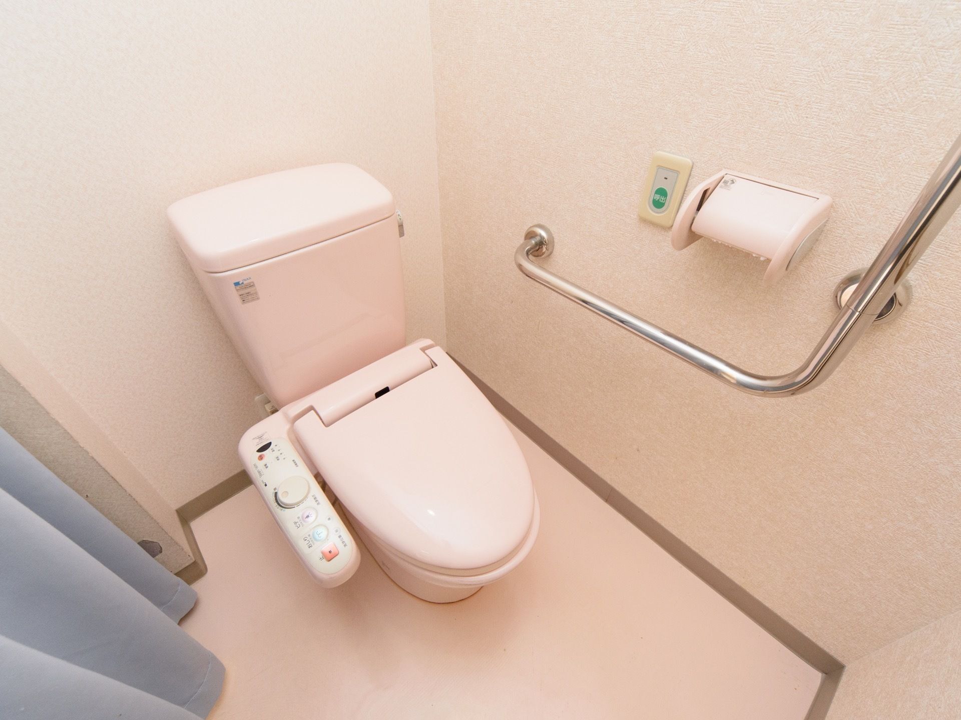 温水洗浄機付きのトイレがひとつある。カーテンで仕切りがされている。壁に手すりとトイレットペーパーホルダー、呼び出しボタンが付いている。