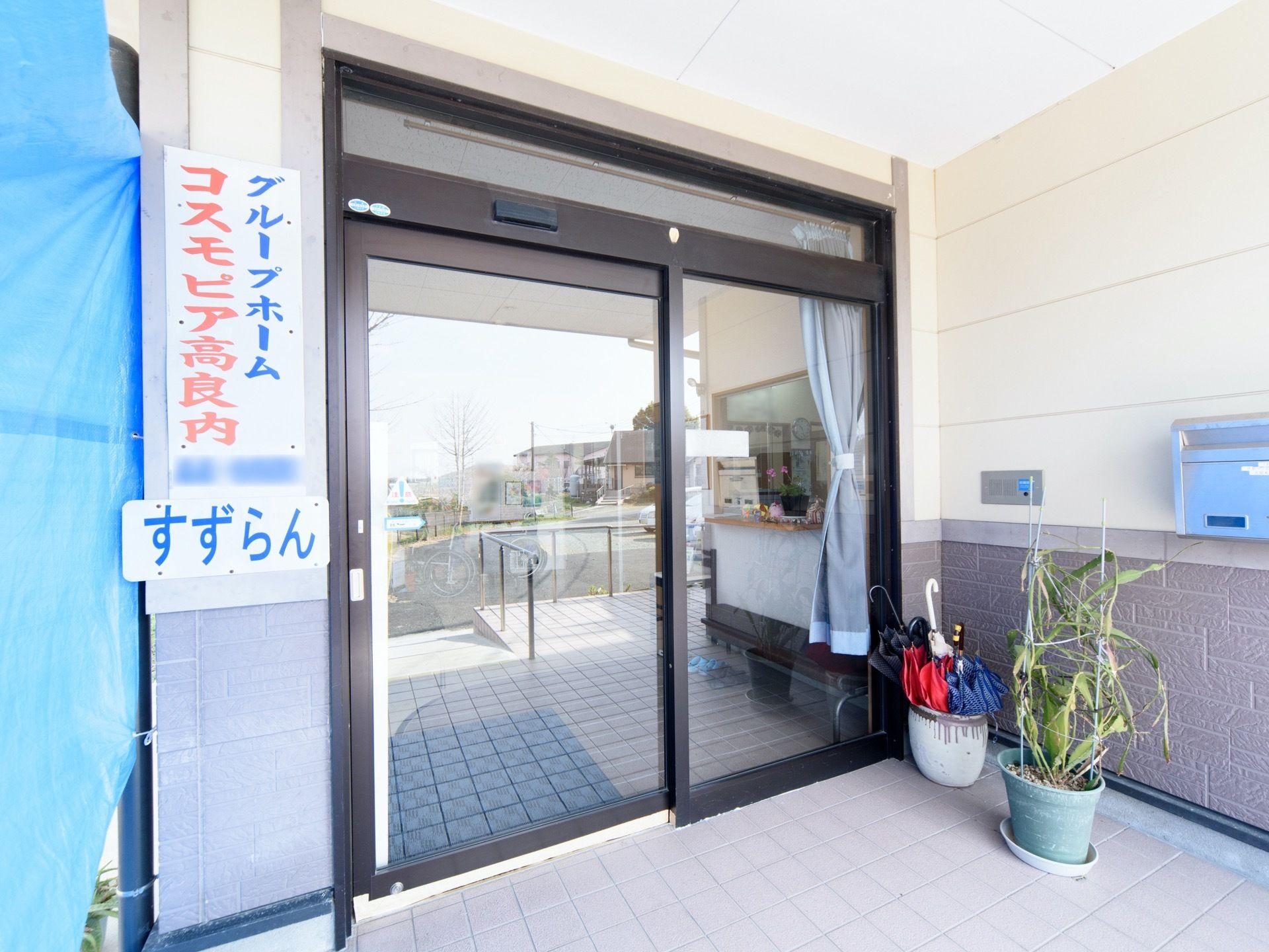 看板の隣に片側開きの自動ドアがある。傘立てと植物が置かれている。看板の隣にはビニールシートが貼られている。