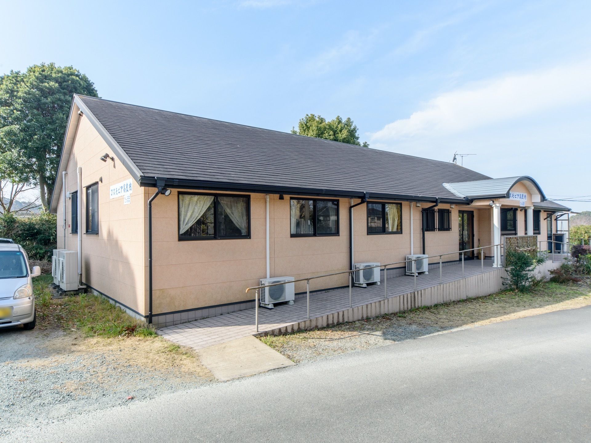道路からスロープがつながっている。隣に駐車場がある。１階建てで三角屋根の建物。壁はベージュで屋根は濃い茶色をしている。
