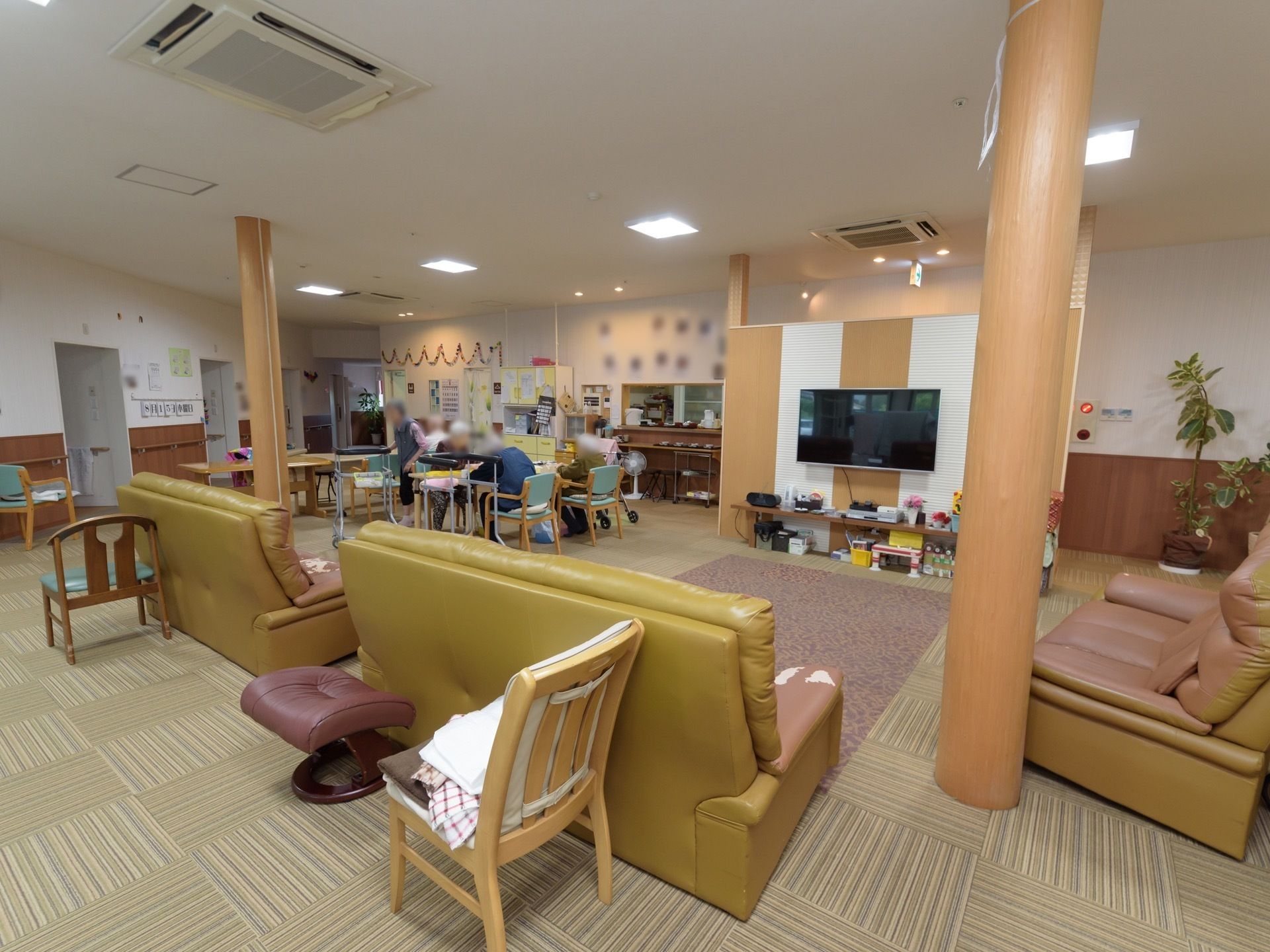 食堂の椅子に座り、入居者様が思い思いの時間を過ごしている。キッチンが併設されており、壁には装飾が施されている。