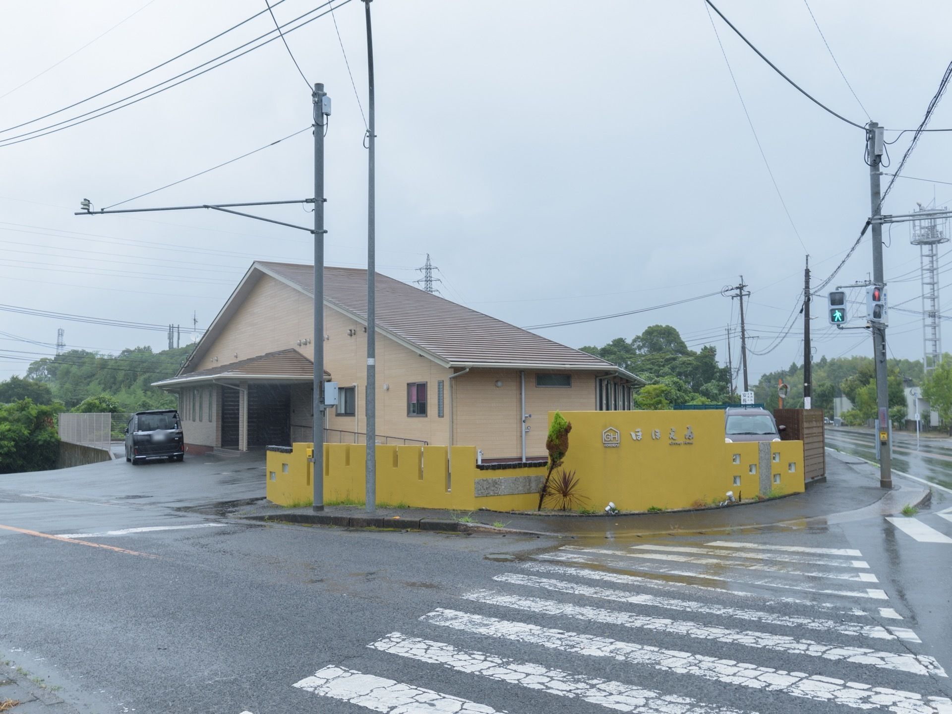 落ち着いた色合いの建物で、玄関横に車が停められている。大きな道路に面しており、目の前には横断歩道が設けられている。