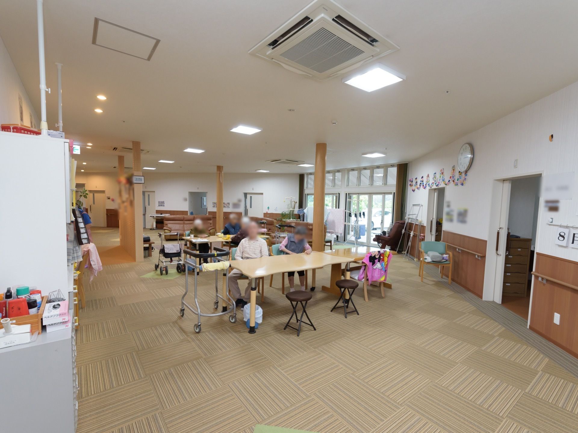 居室前に設けられている食堂で、複数の入居者様が椅子に座って寛いでいる。大きな窓からは駐車場が見える。