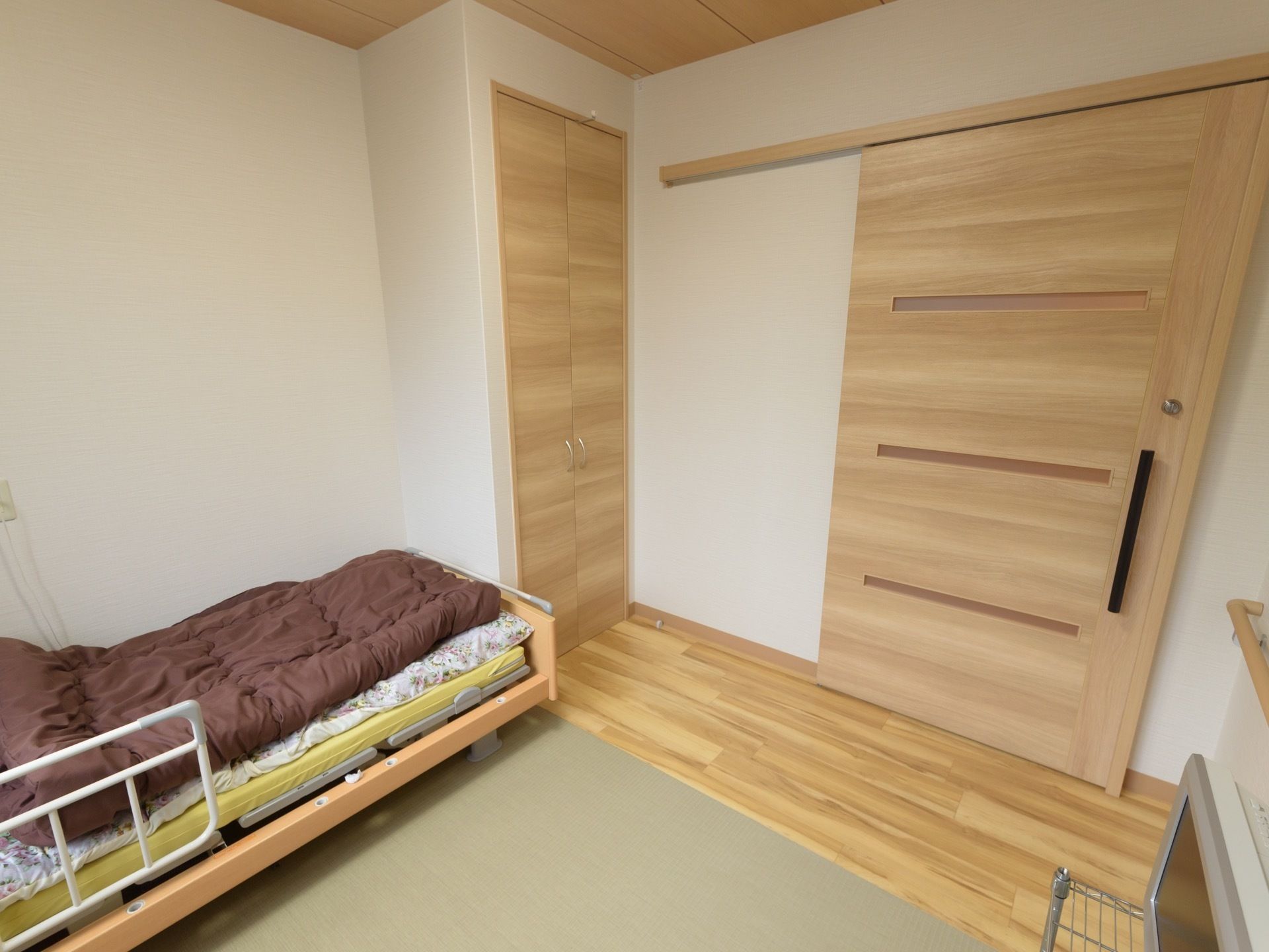 木製の引き戸が付いた居室がある。壁際には介護用ベッドが置かれていて、足元には備え付けの収納が付いている。