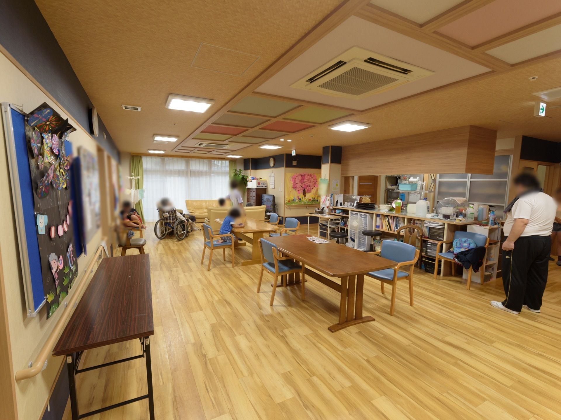 テーブルや椅子がたくさん並べられたフロアがあり、入居者様がくつろいでいる。天井には空調がある。