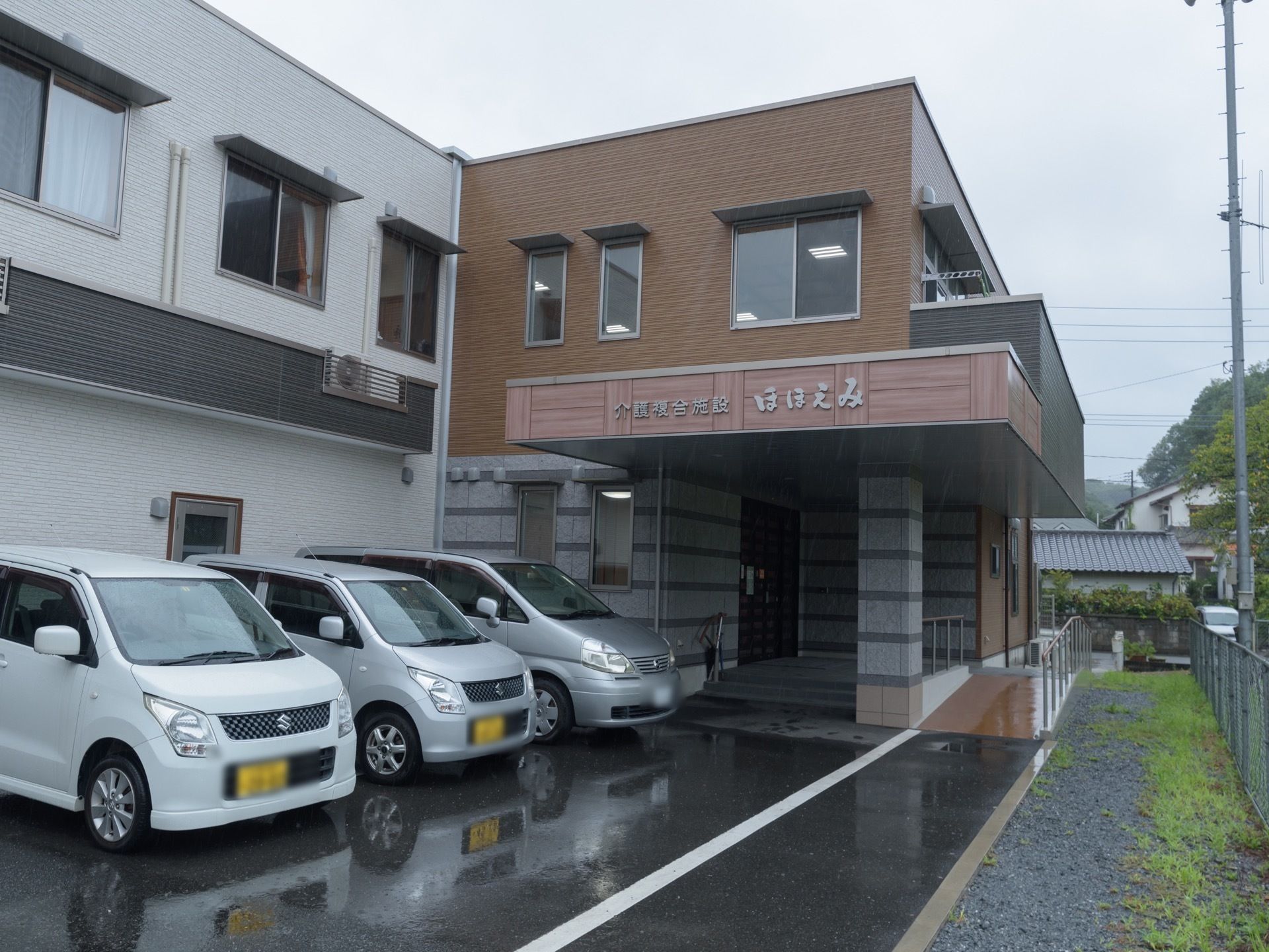 ２階建ての建物で、玄関には階段とスロープがある。敷地内には駐車場があり、複数台の車が停められている。