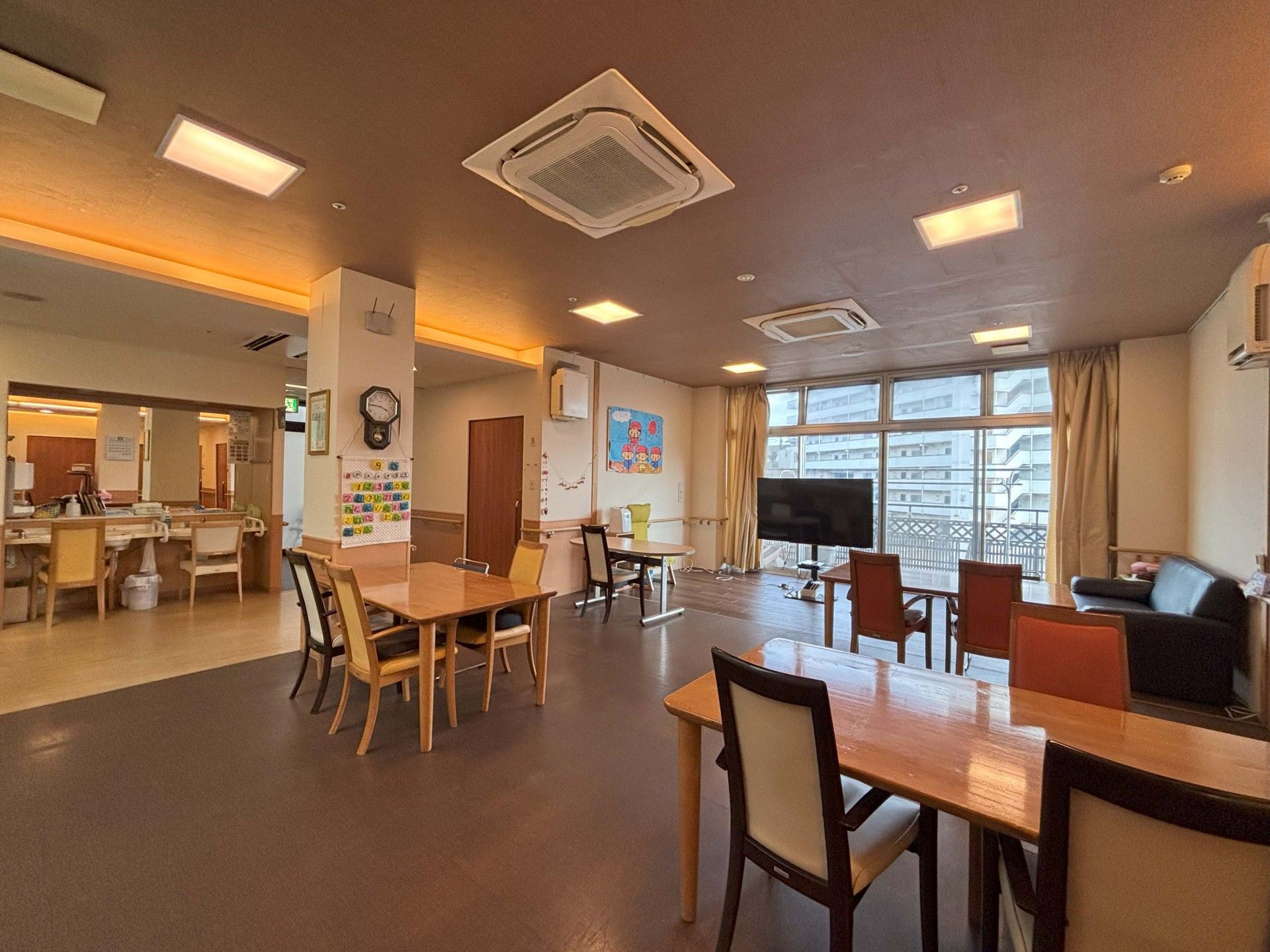 ブルーのポロシャツを着た女性スタッフに付き添われて外出を楽しんでいる眼鏡をかけた車いすの高齢女性。周辺には緑が多くある。