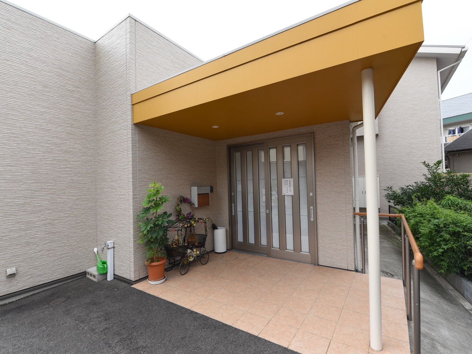 黄色い屋根付きのエントランスである。手すりが右側に設置されている。壁際には、観葉植物や傘立てが置かれている。
