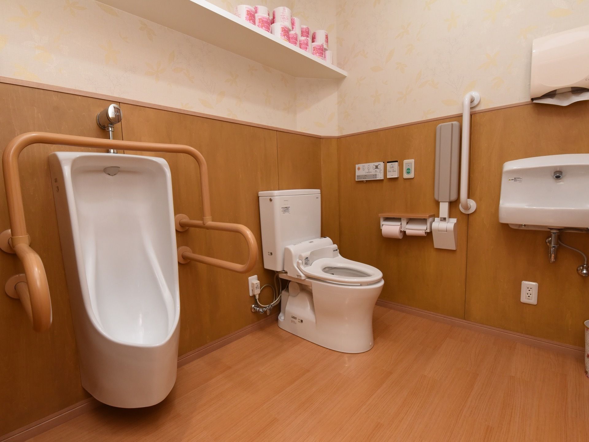 男性用小便器と洋式便器が設置された広いトイレである。洗面台やペーパータオルホルダーが備わっている。手すりが壁に取りつけられている。