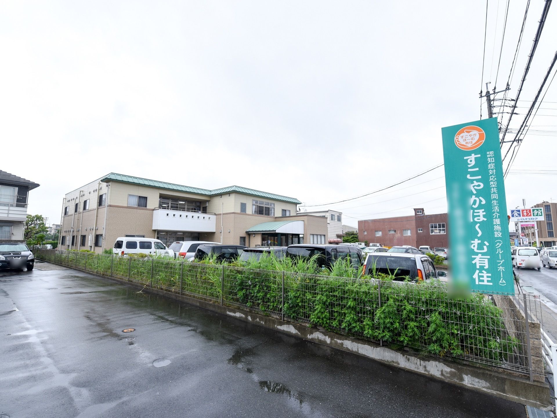 大きなブラウン色の建物。入り口に施設名の書かれた緑色の看板が立っており、駐車スペースが用意されている。
